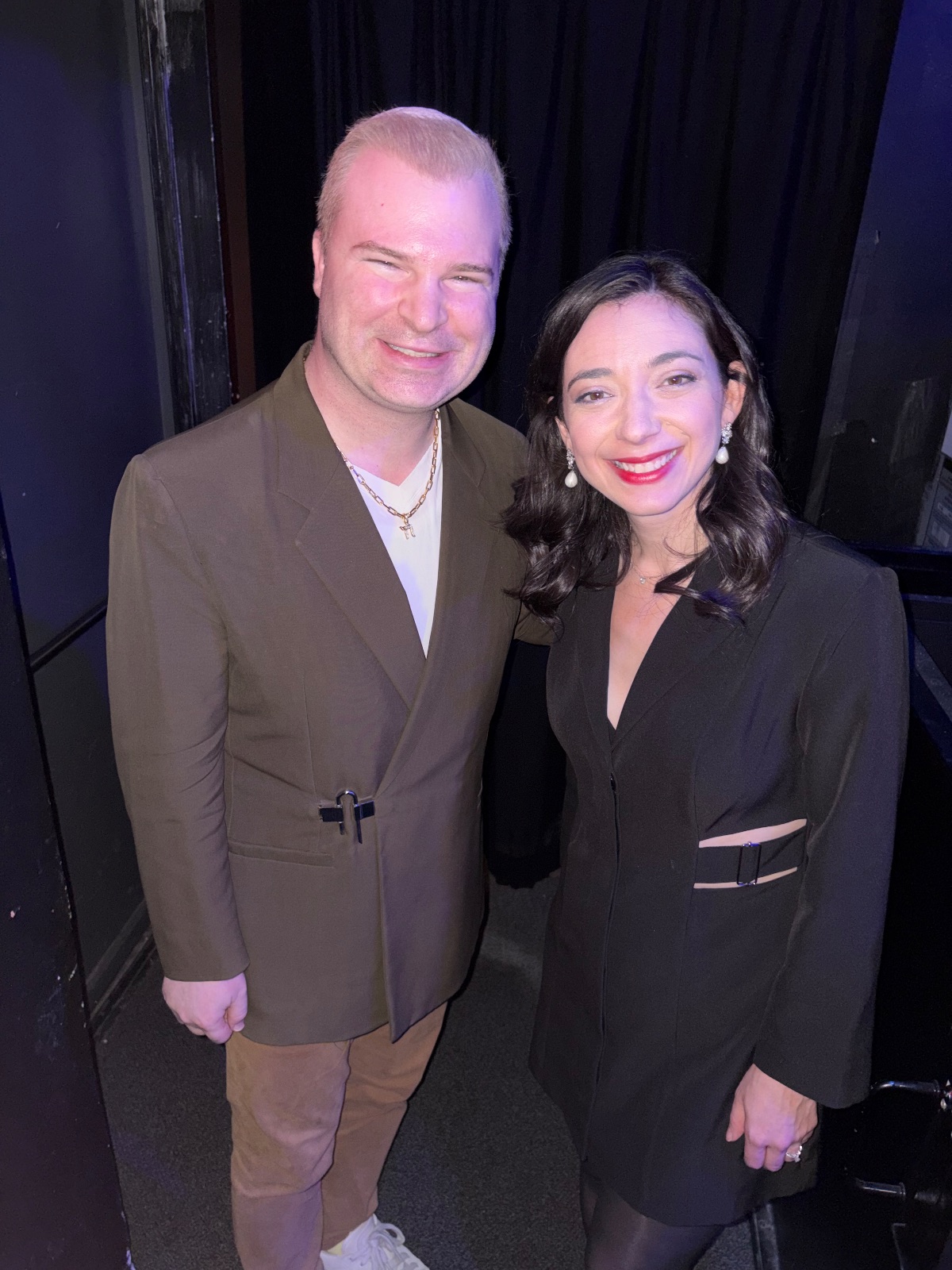 Two people in formal attire smiling together in a warm backstage setting.