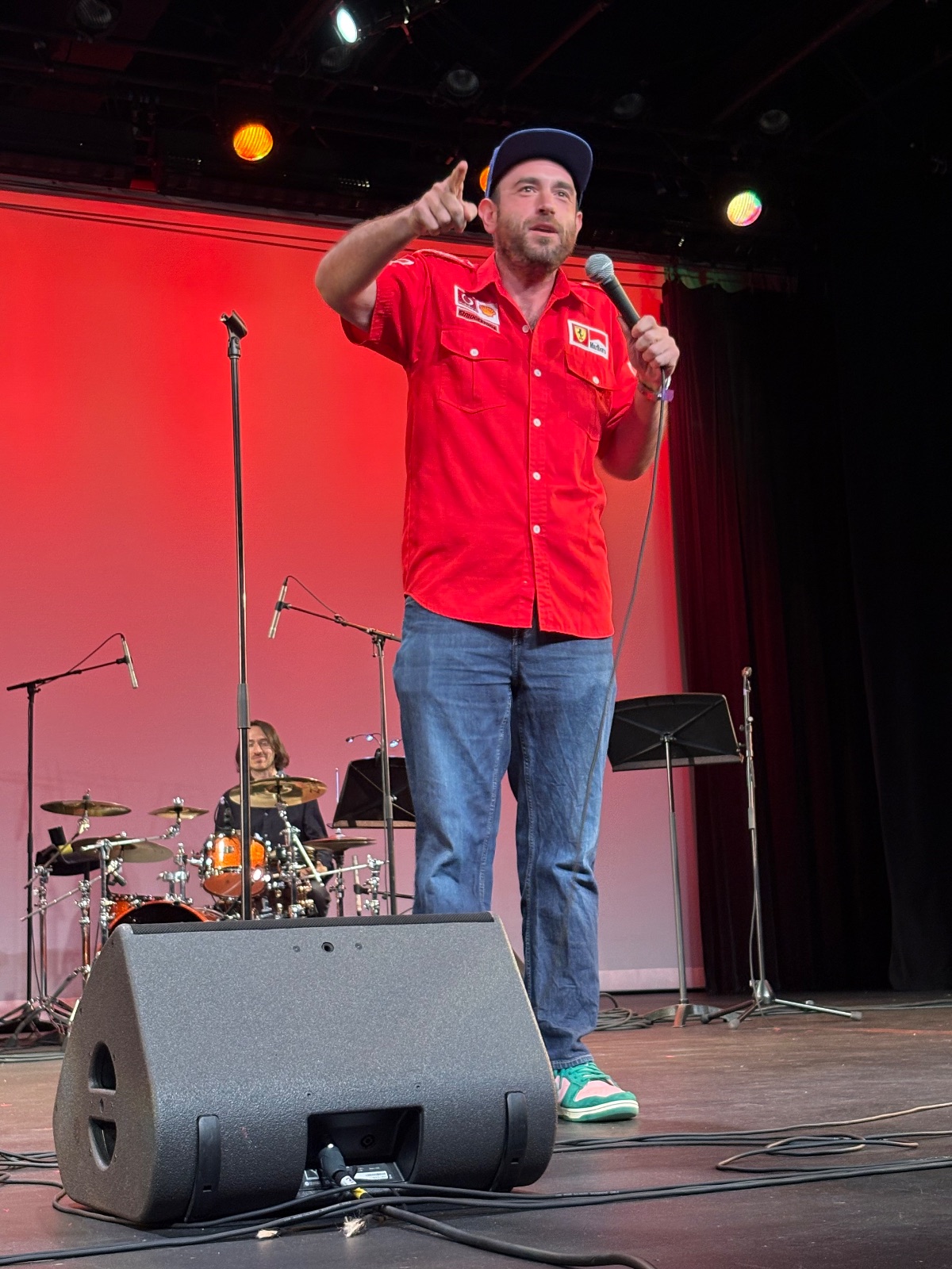 A performer on stage with raised arms gestures dynamically toward the audience under professional lighting against a red back