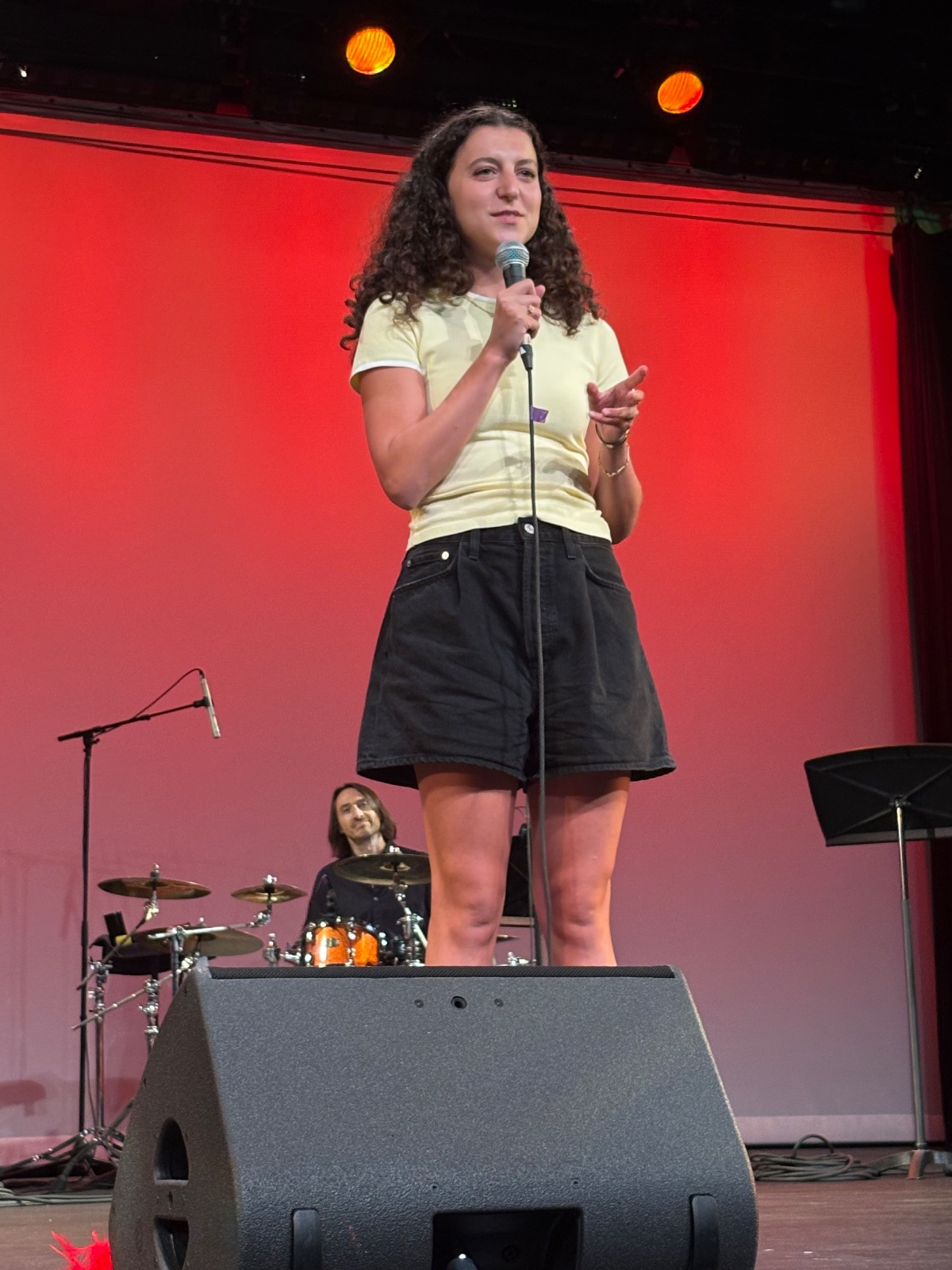 A performer stands on stage under red lighting, facing the audience with confident posture.