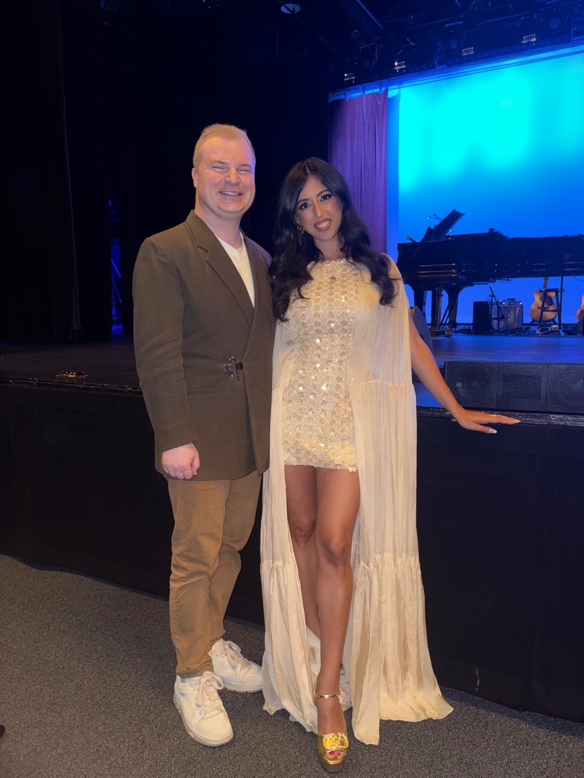 Two people in formal attire stand on stage near a grand piano with blue atmospheric lighting.