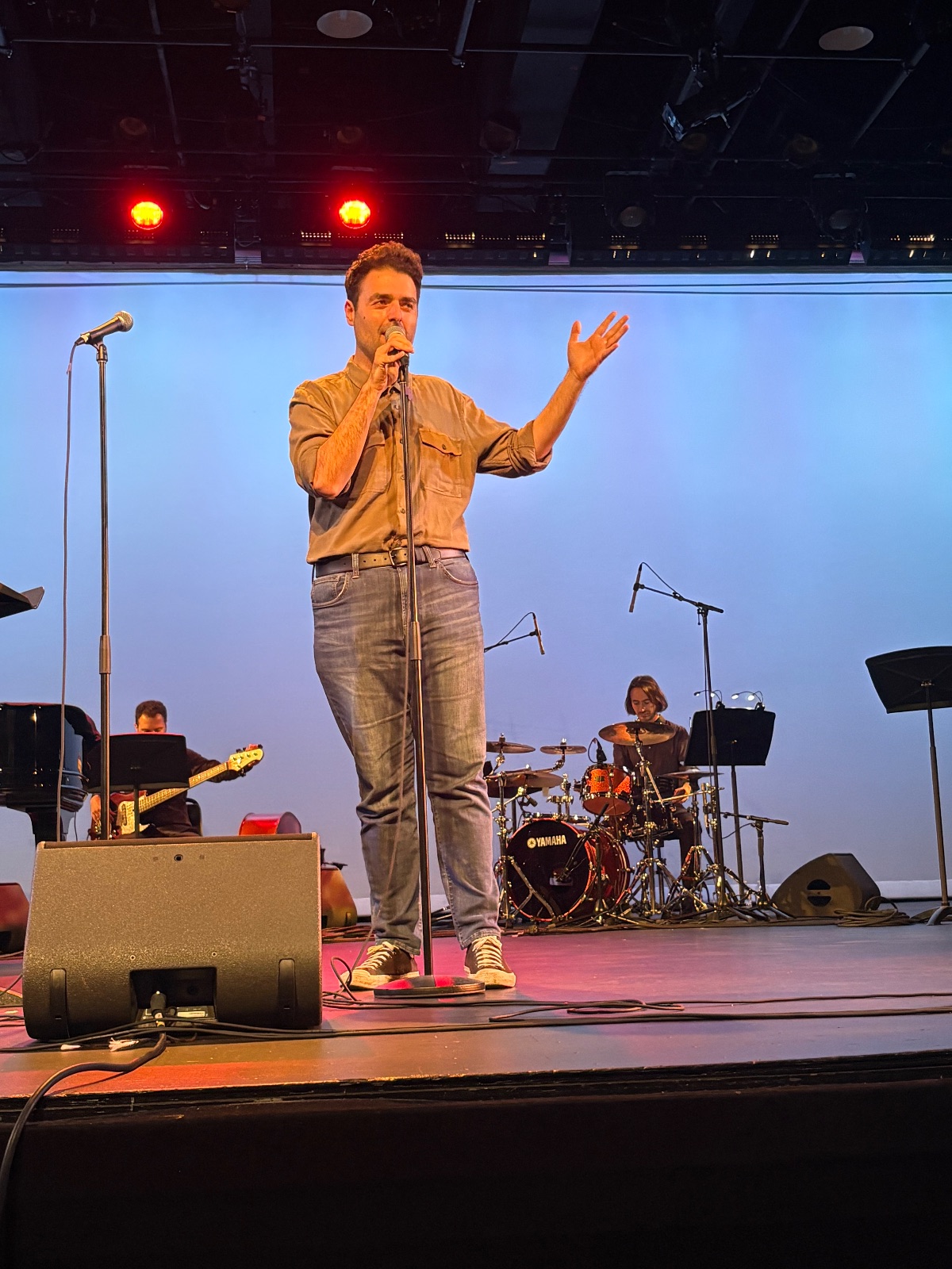 A performer stands on stage singing into a microphone with presence and expression.
