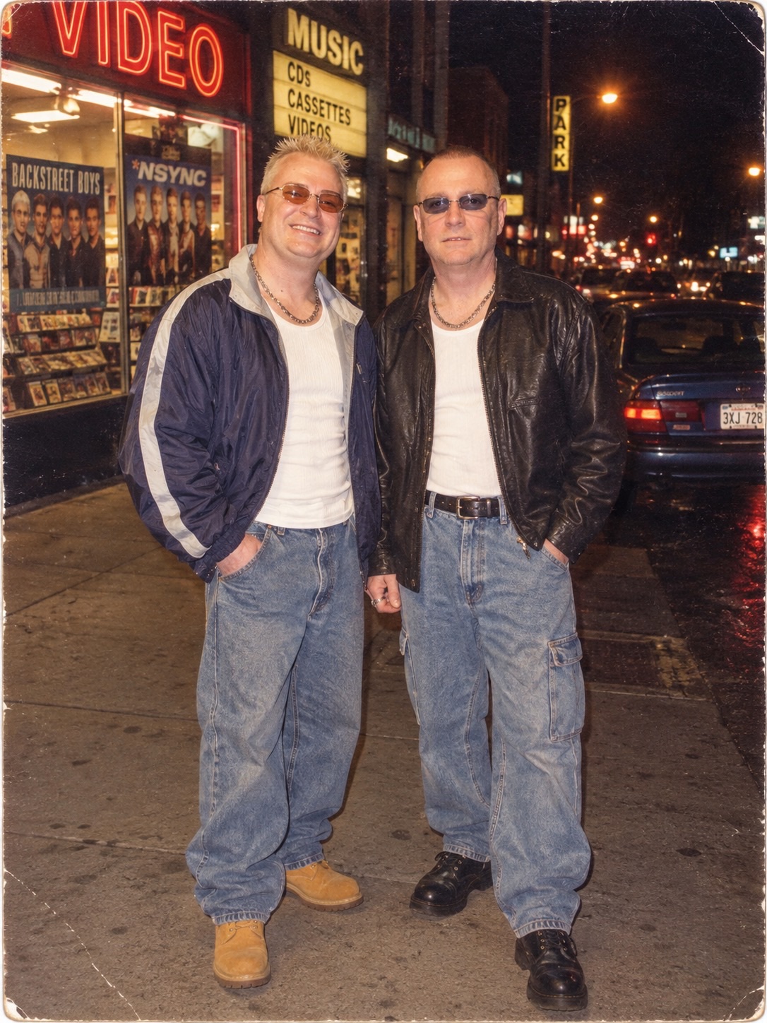 Two people stand outside a video store on a city street at night.